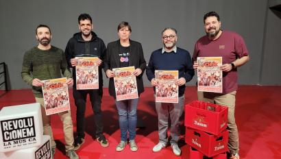 Cinc persones posen davant d’un fons neutre sostenint cartells promocionals del Super Folk Festival. A l’esquerra, dos cubs amb el text “Revolucionem la música”. Al terra, caixes vermelles amb el logotip de Damm. Tots somriuen i mostren els cartells de l’esdeveniment.